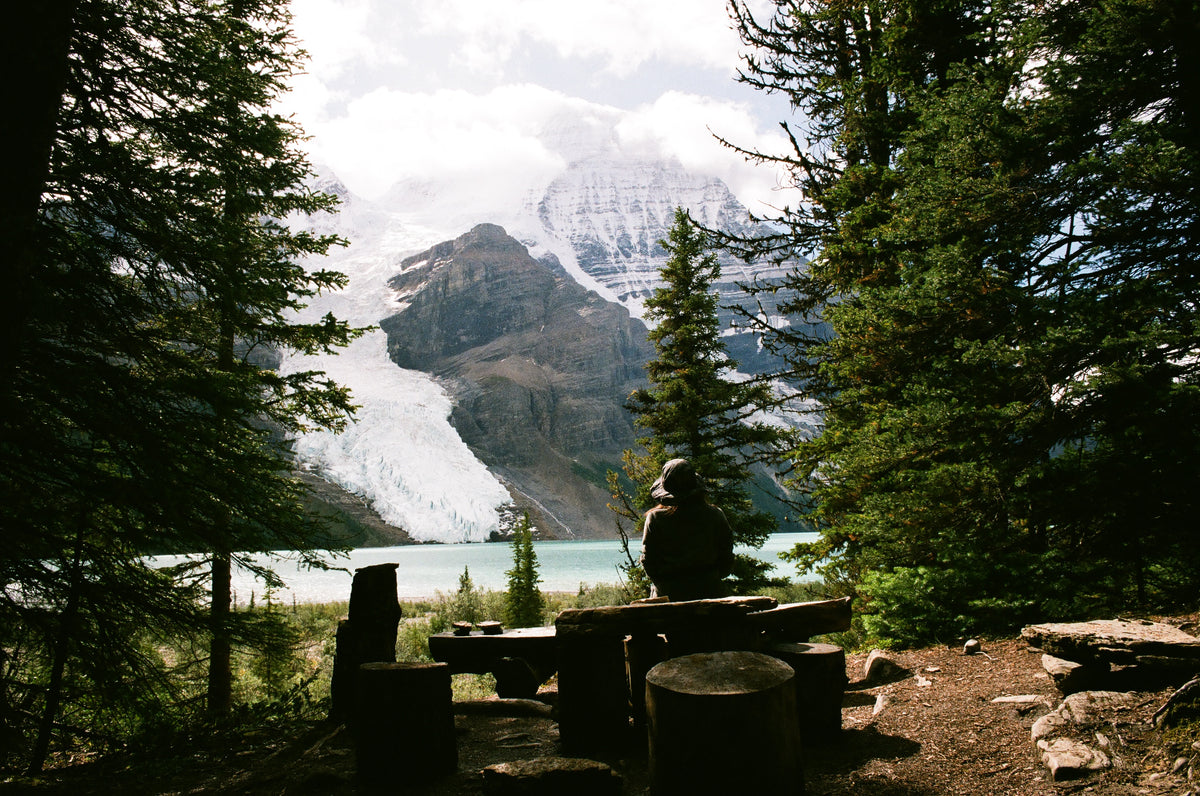 Hiker resting beside a turquoise mountain lake framed by forest and snow-capped peaks
